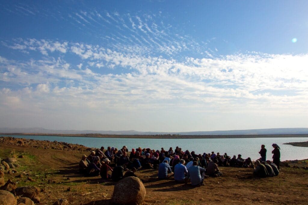 A group of about 40-50 adults sit in a loose circle, facing 2 people who are standing, presenting to the group. The group is seated on a dirt clearing next to a bright blue body of water, with clouds streaking the sky and rolling hills in the distance.