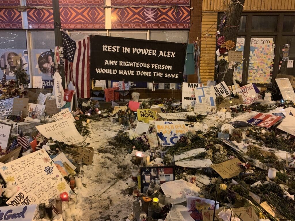 Brightly colored memorial site for Alex Pretti. Flowers and posters and offerings adorn a snow bank in front of a large banner that reads "Rest in Power Alex. Any righteous person would have done the same." Next to the banner hangs an American flag in honor of the martyred ICU nurse who cared for veterans in Minnesota.