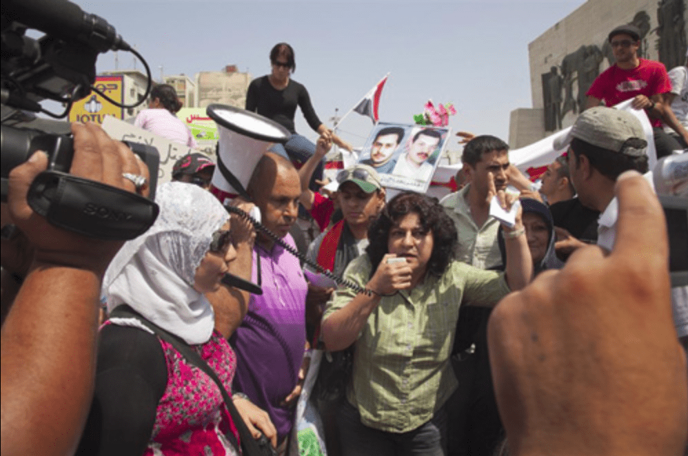 A brightly colored photo of protestors and media surrounds a young Yanar Mohammed speaking into a megaphone in Iraq